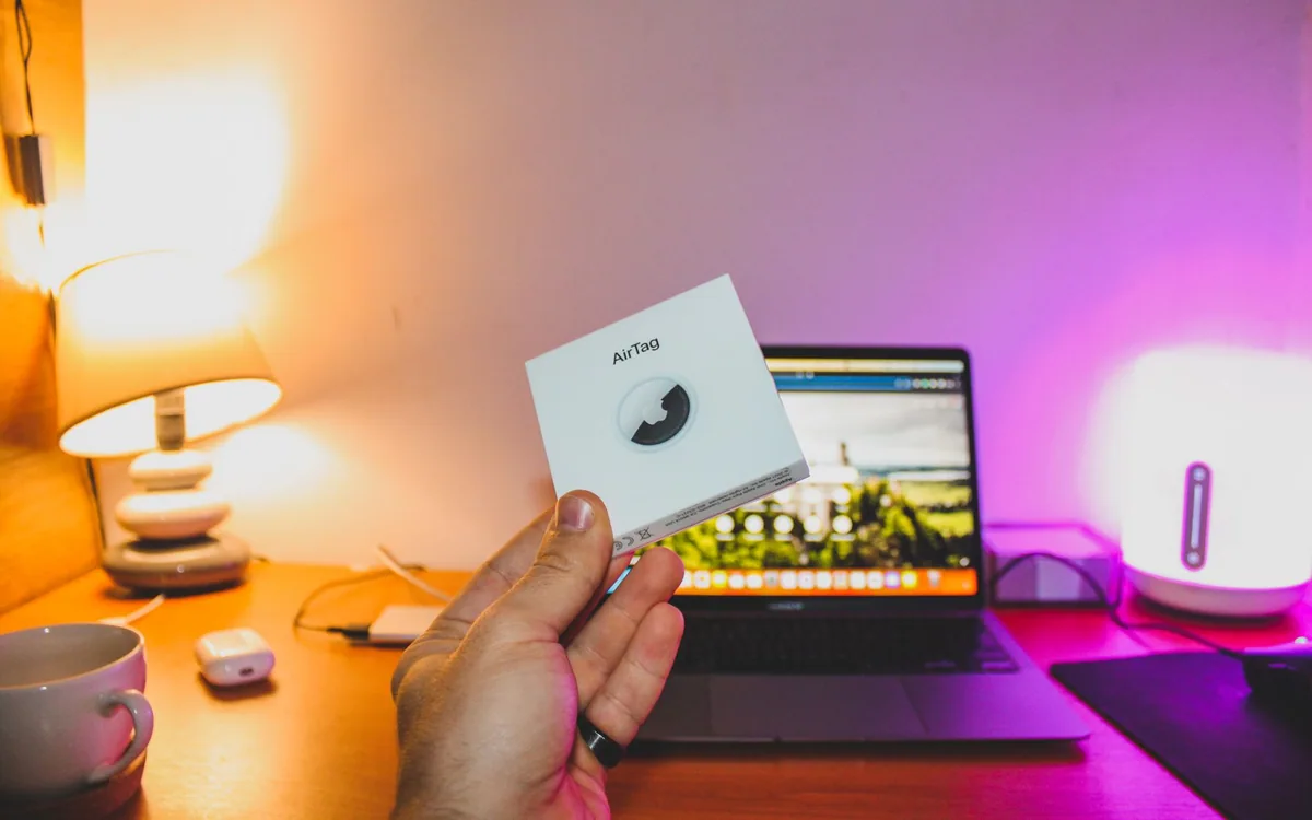 A hand holds an AirTag box in front of a laptop on a home desk.