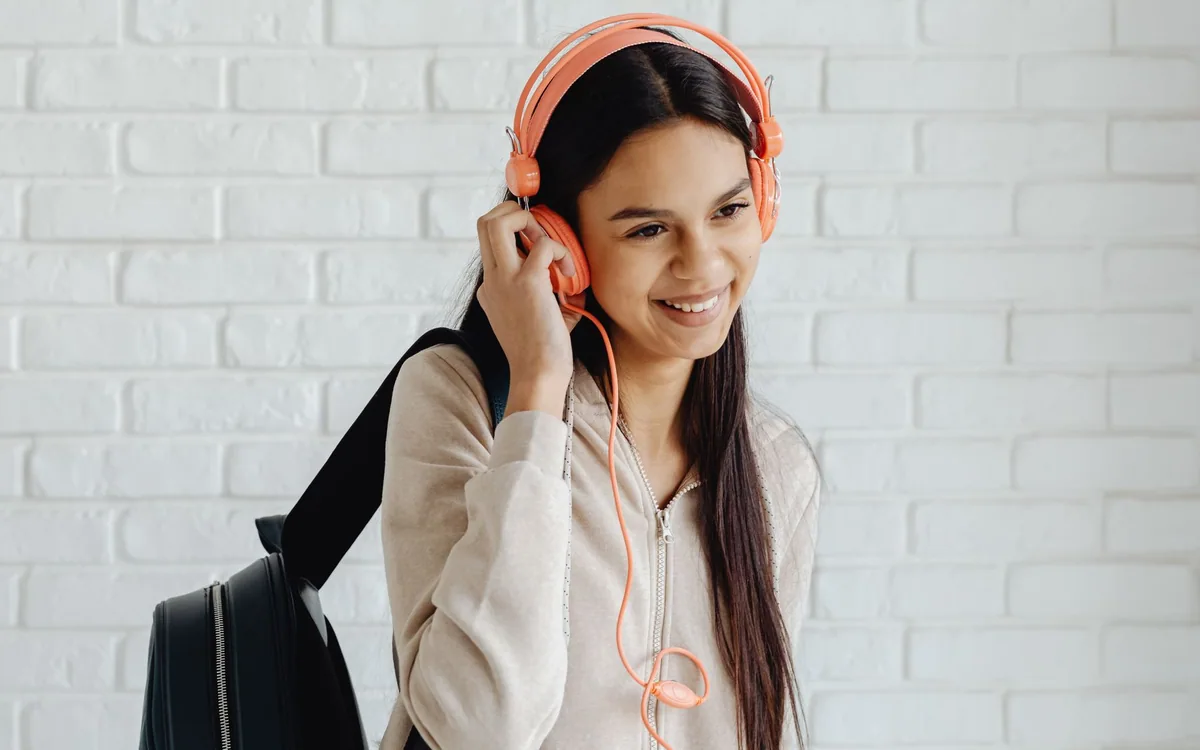 A smiling young woman wears pink wired headphones and holds a backpack.